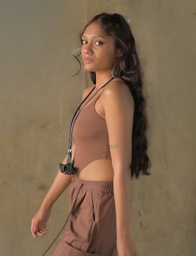 Young woman with long dark hair wearing brown cut-out top and pants, black sunglasses hanging on necklace