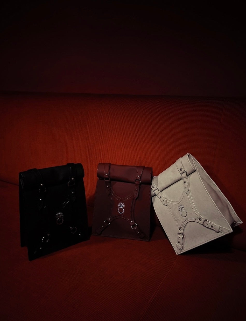 Three leather roll-top backpacks in black, brown, and white on a red fabric background