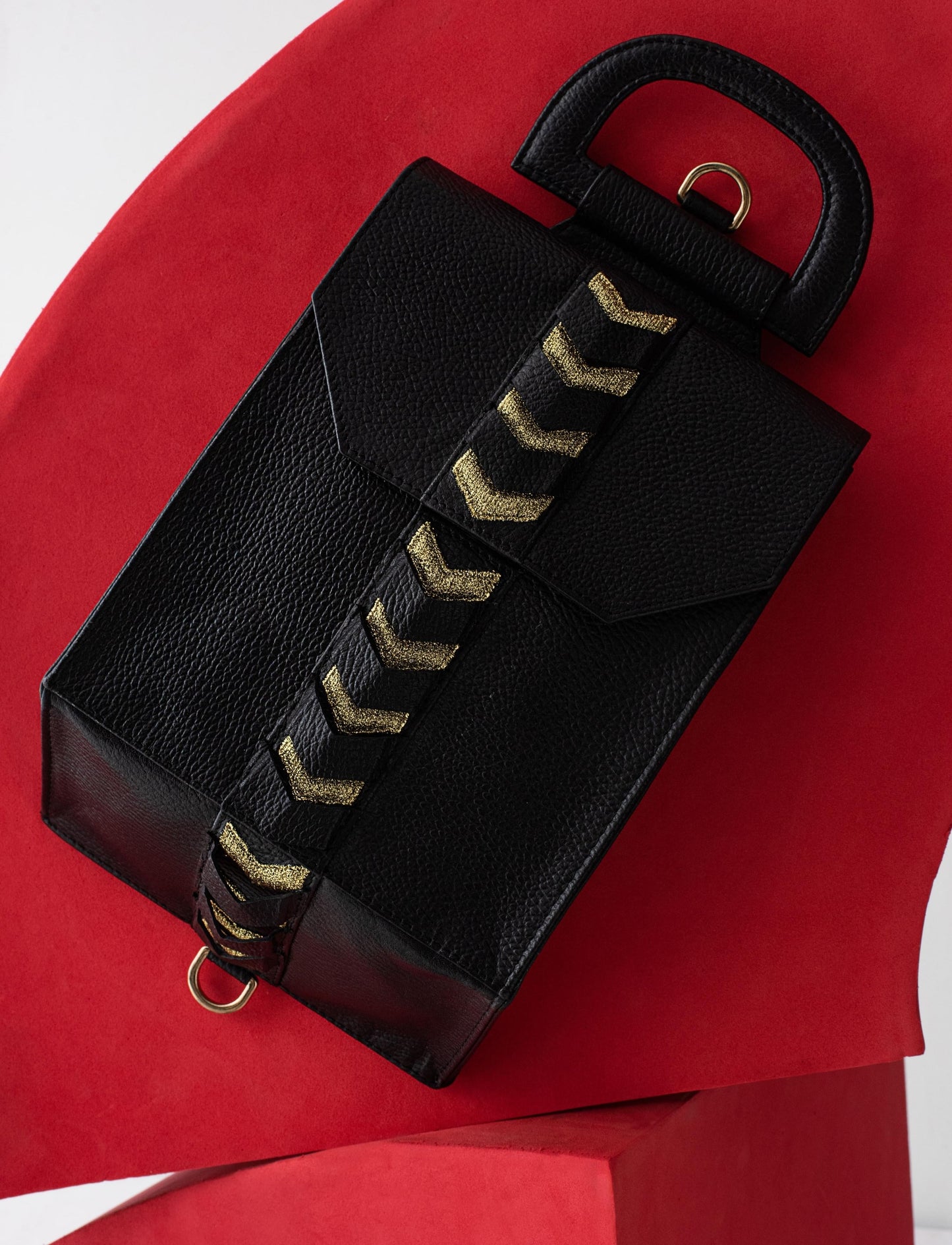 Black textured leather handbag with gold chevron strap on curved red surface