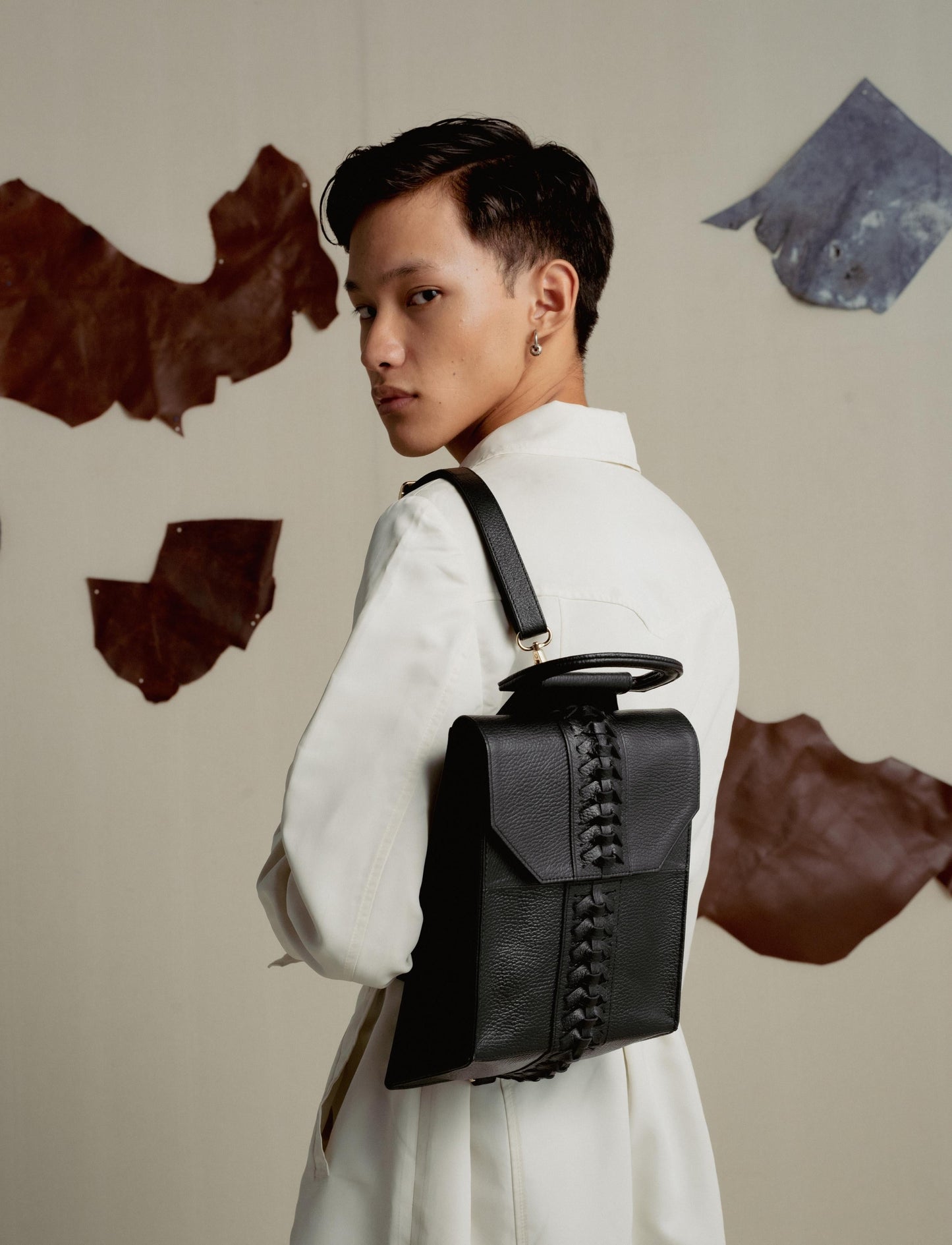 Young person in white jacket with black leather backpack featuring woven detail, neutral background with leather patches