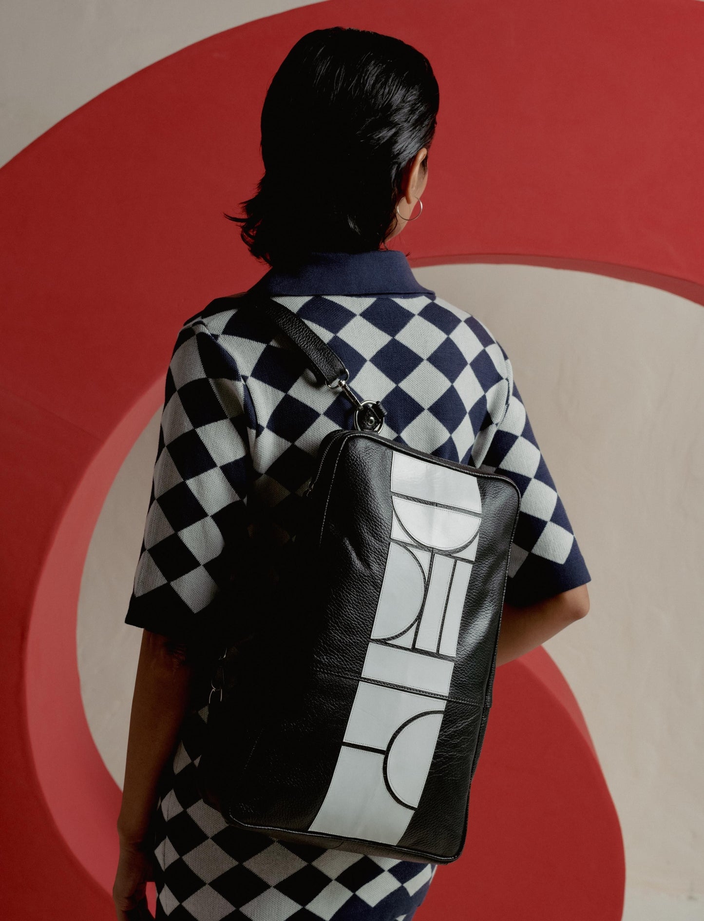 Person with short dark hair wearing black and white checkered shirt and black leather bag with white geometric design