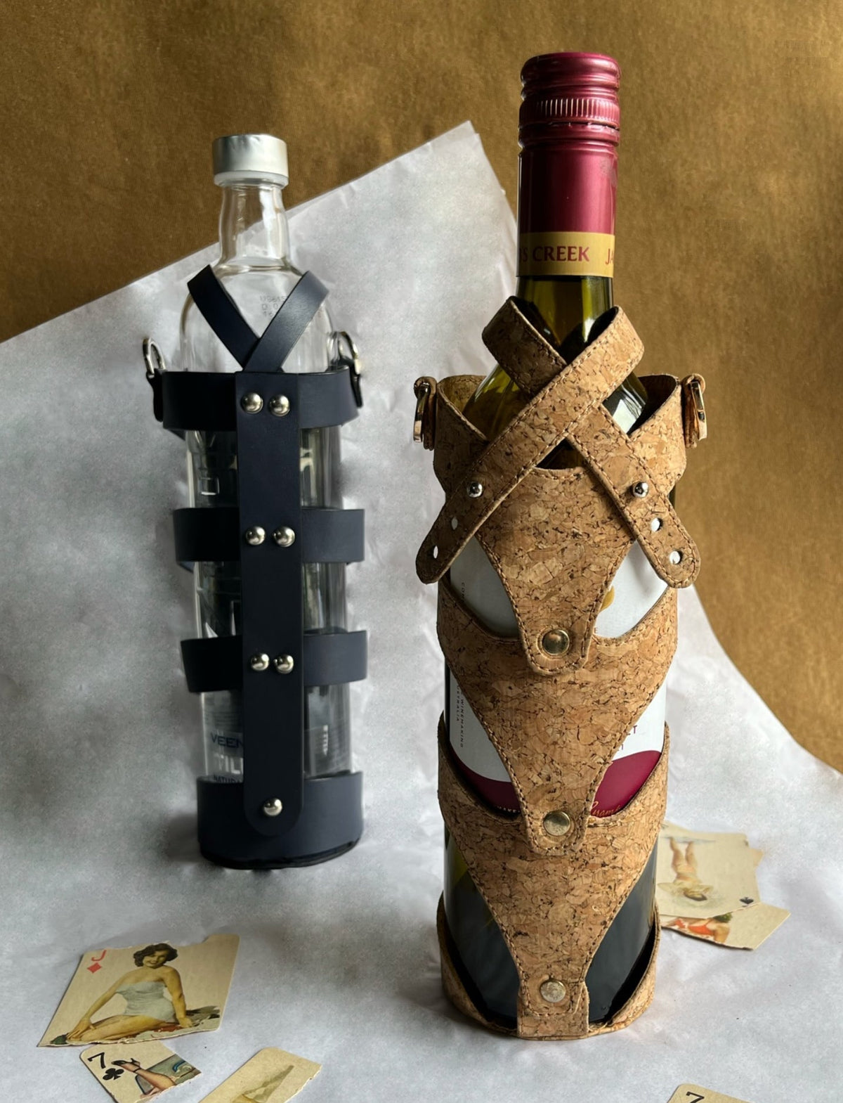 Two bottles, one wine and one clear, in stylish cork and black leather upcycled stitched wine cases on white surface with vintage playing cards