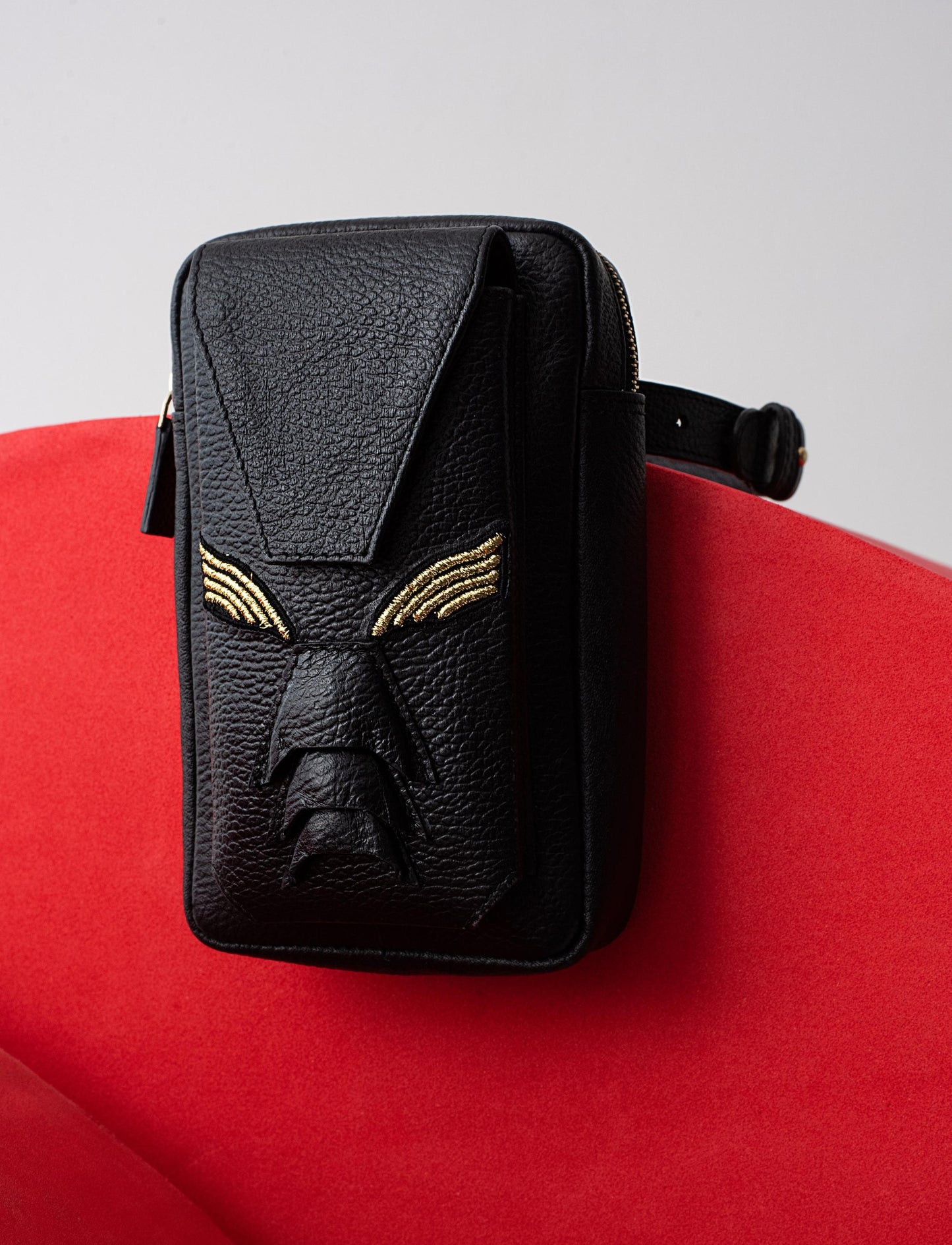 Black leather crossbody bag with embossed face design and gold accents on red chair