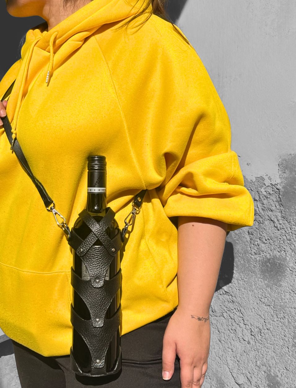 Person wearing bright yellow hoodie carrying black leather upcycled stitched wine bottle case with adjustable strap