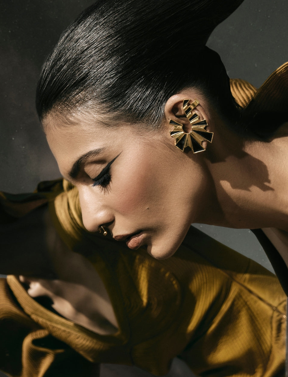 Close-up side profile of woman with sleek black hair wearing large geometric gold earrings and mustard yellow garment