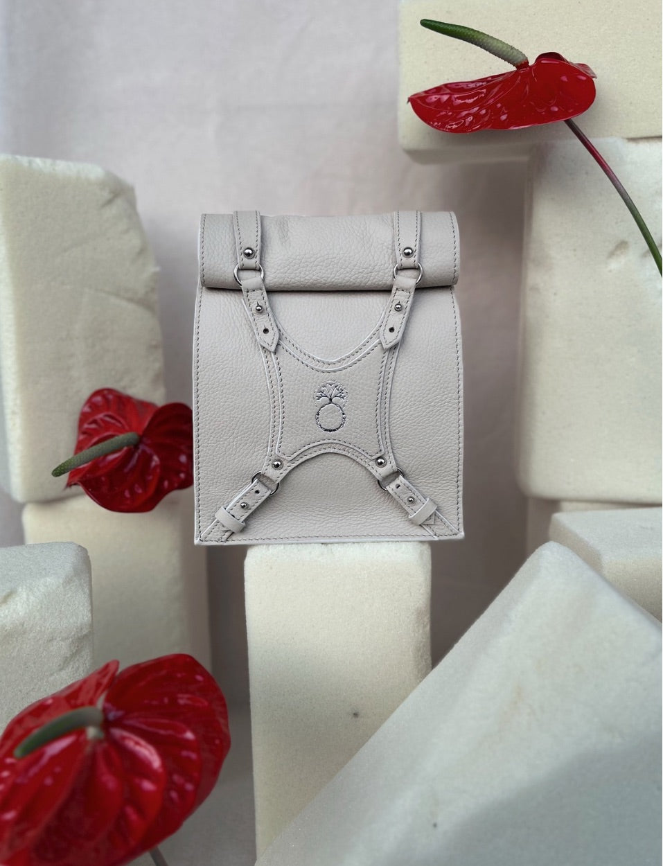 Minimalist beige leather backpack with decorative straps, surrounded by red anthurium flowers and foam blocks