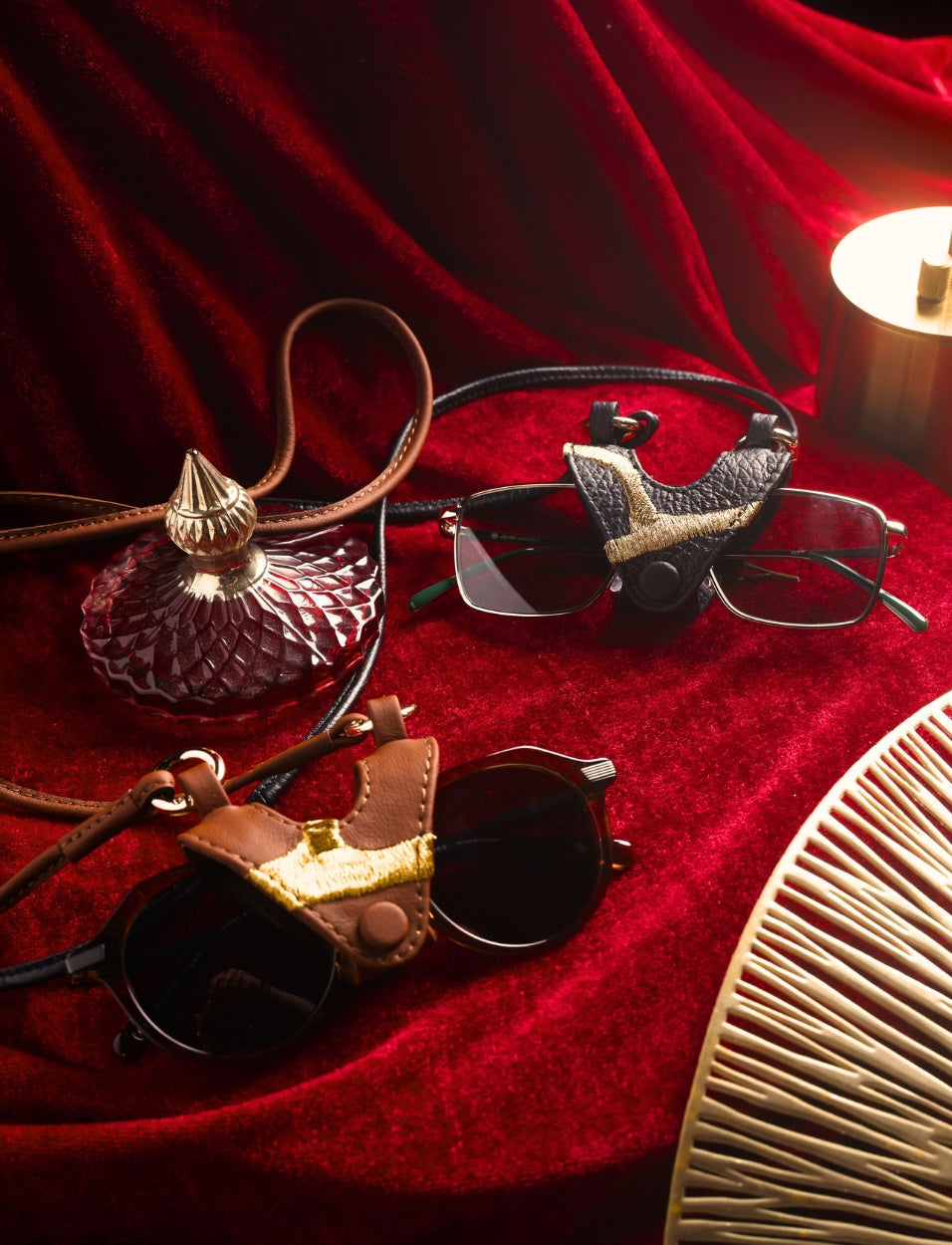 Two pairs of glasses with leather holders on textured red velvet, next to a vintage glass perfume bottle and a brass lamp