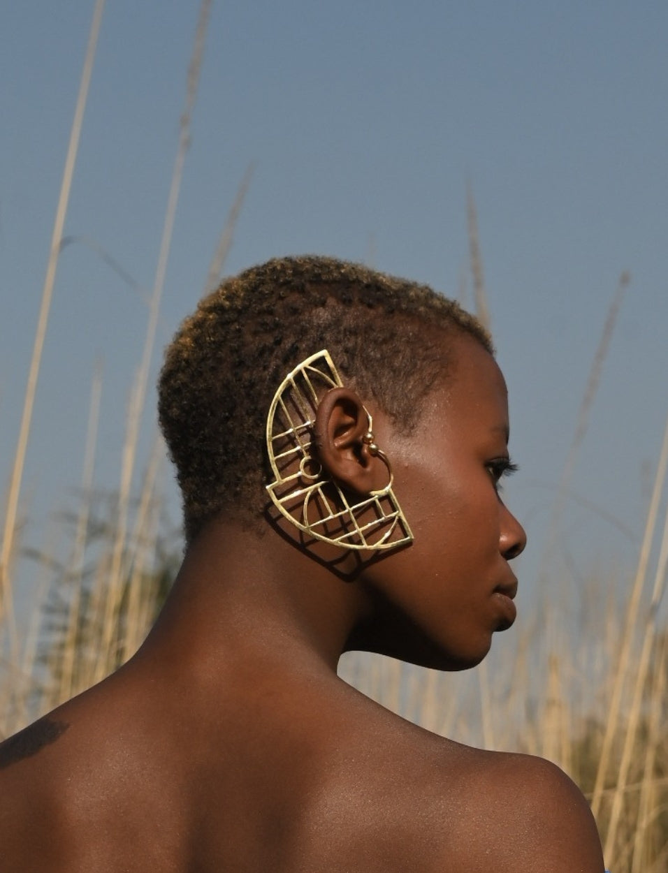 Profile of a woman with short hair wearing large geometric gold earrings outdoors with tall grass and clear blue sky