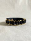 Black leather wristband with gold metal rings woven around it on a beige fabric surface
