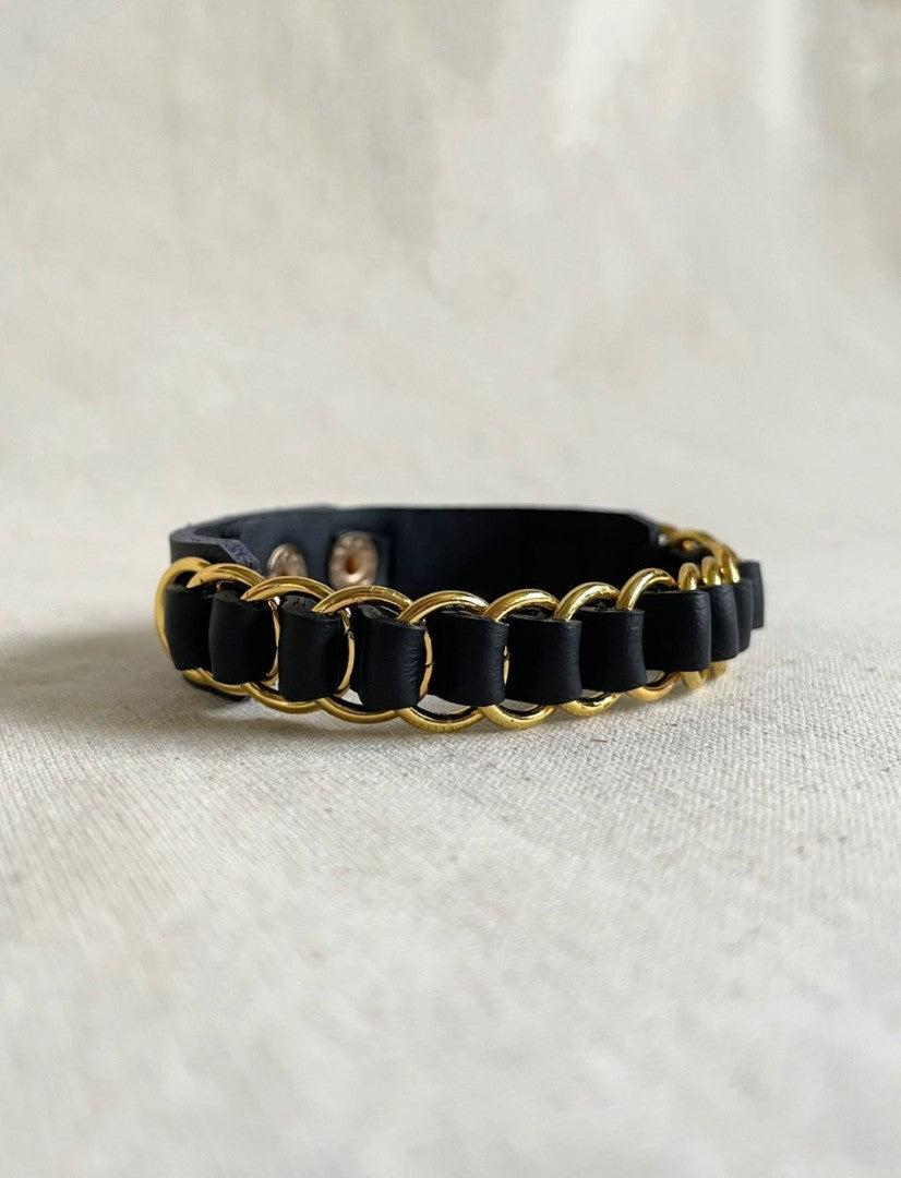 Black leather wristband with gold metal rings woven around it on a beige fabric surface