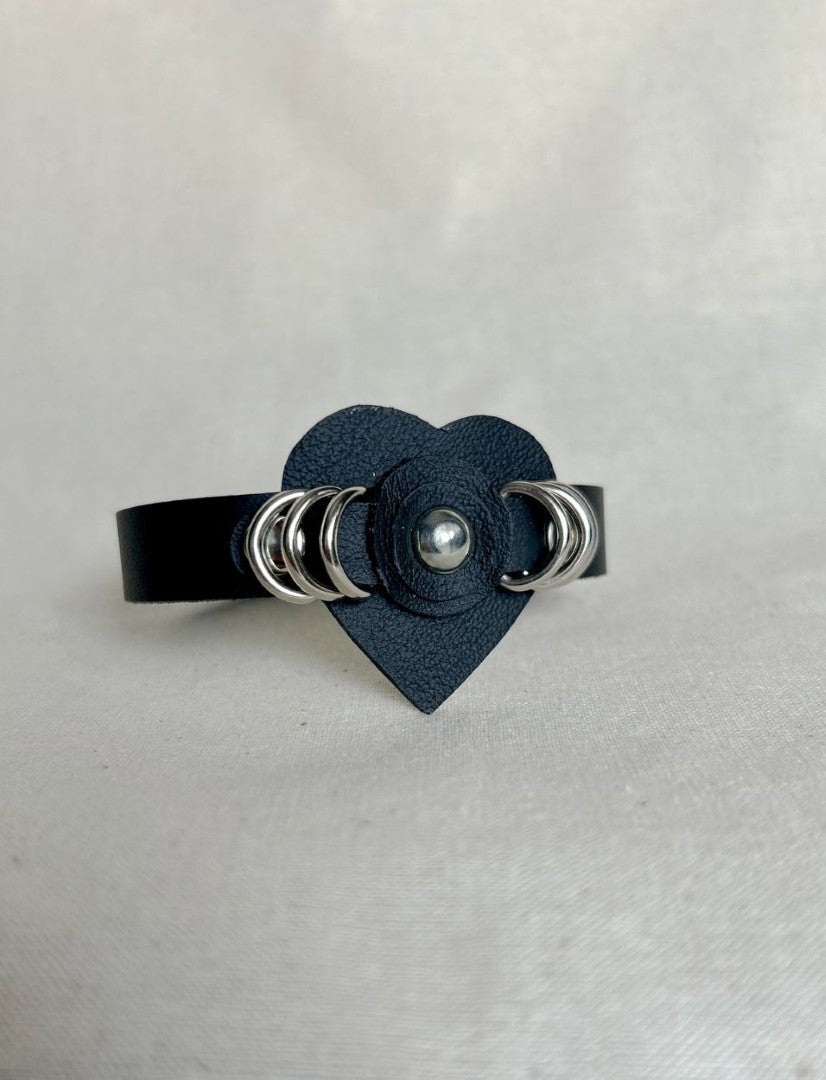 Black leather cuff bracelet with heart-shaped centerpiece and silver ring studs on neutral background