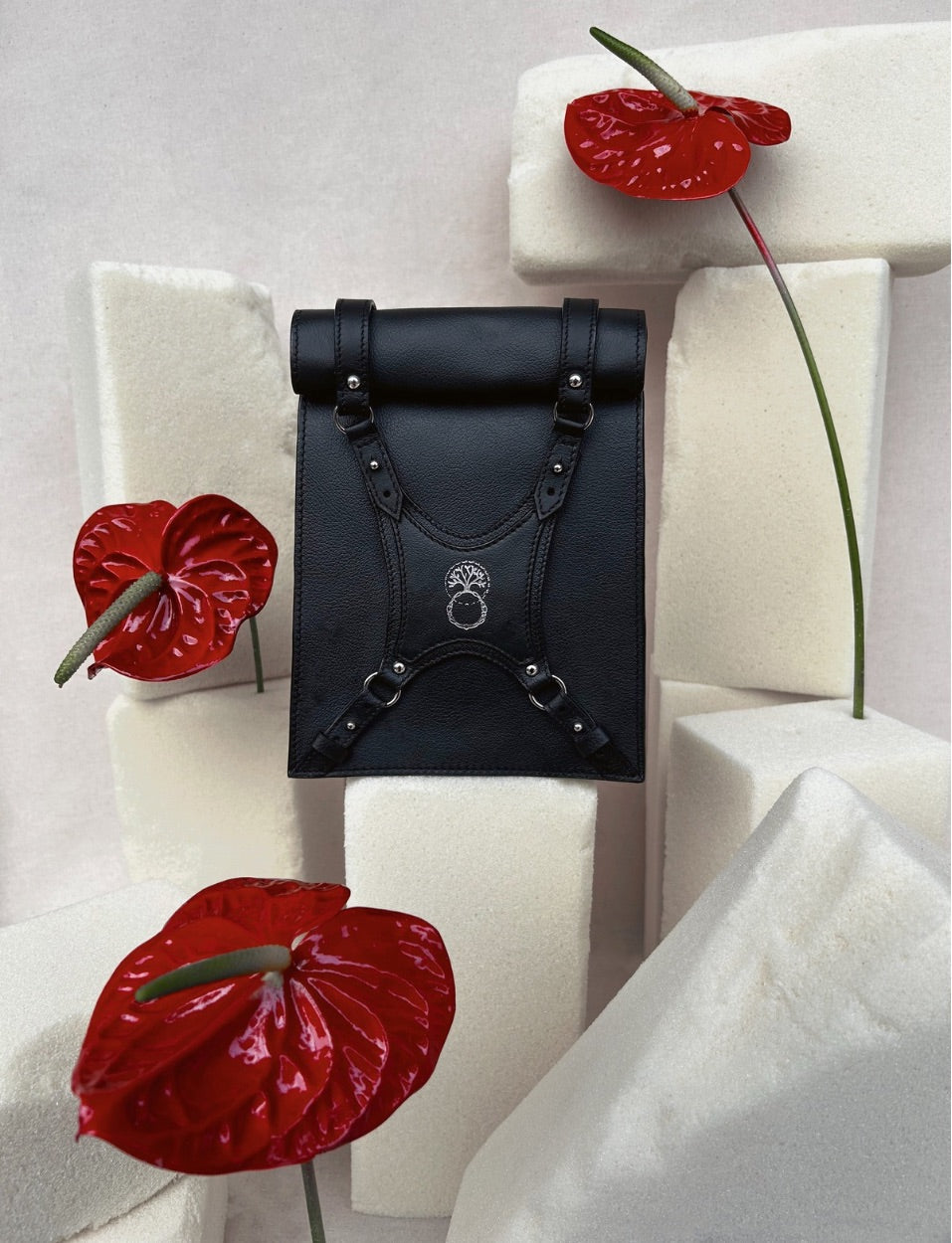 Black leather bag with straps and emblem, surrounded by red anthurium flowers on white foam blocks