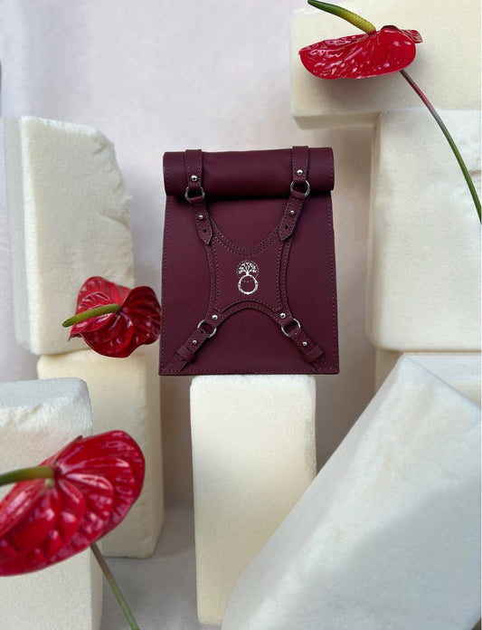 Maroon leather roll-top backpack with silver buckles, displayed on white foam blocks with red anthurium flowers