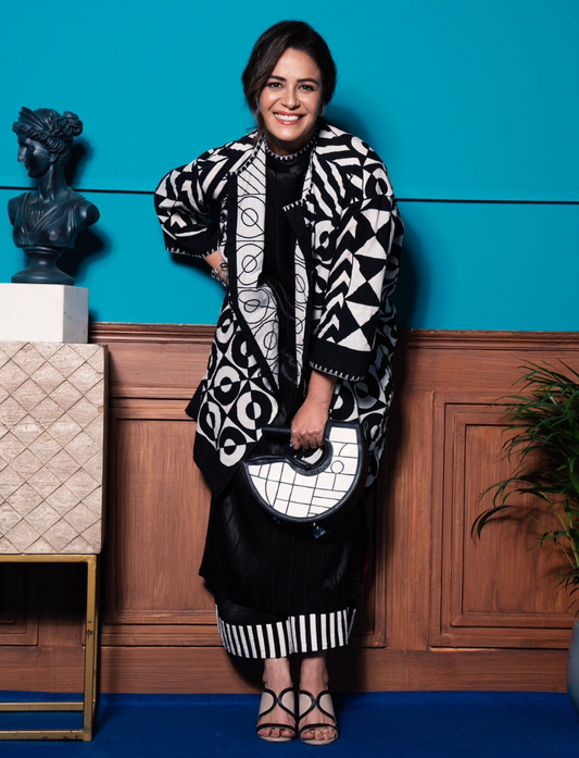 Smiling woman in black and white geometric print outfit holding matching handbag, standing by blue wall