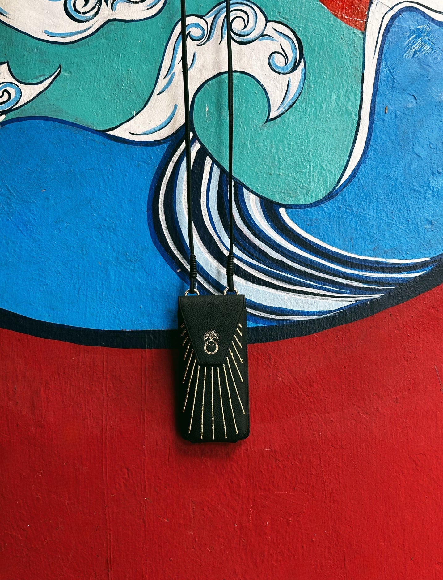 Black leather phone sling bag with gold embroidery hanging against colorful blue, green, red wall mural