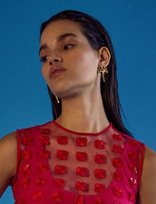 Woman wearing gold sunburst earrings and a red sleeveless top with floral appliqué against a blue background