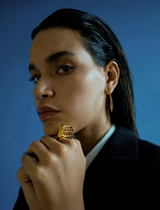 Close-up of woman wearing geometric gold statement ring and matching earrings against blue background