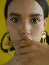Close-up of woman with dark hair wearing gold geometric statement ring and matching earrings against yellow background