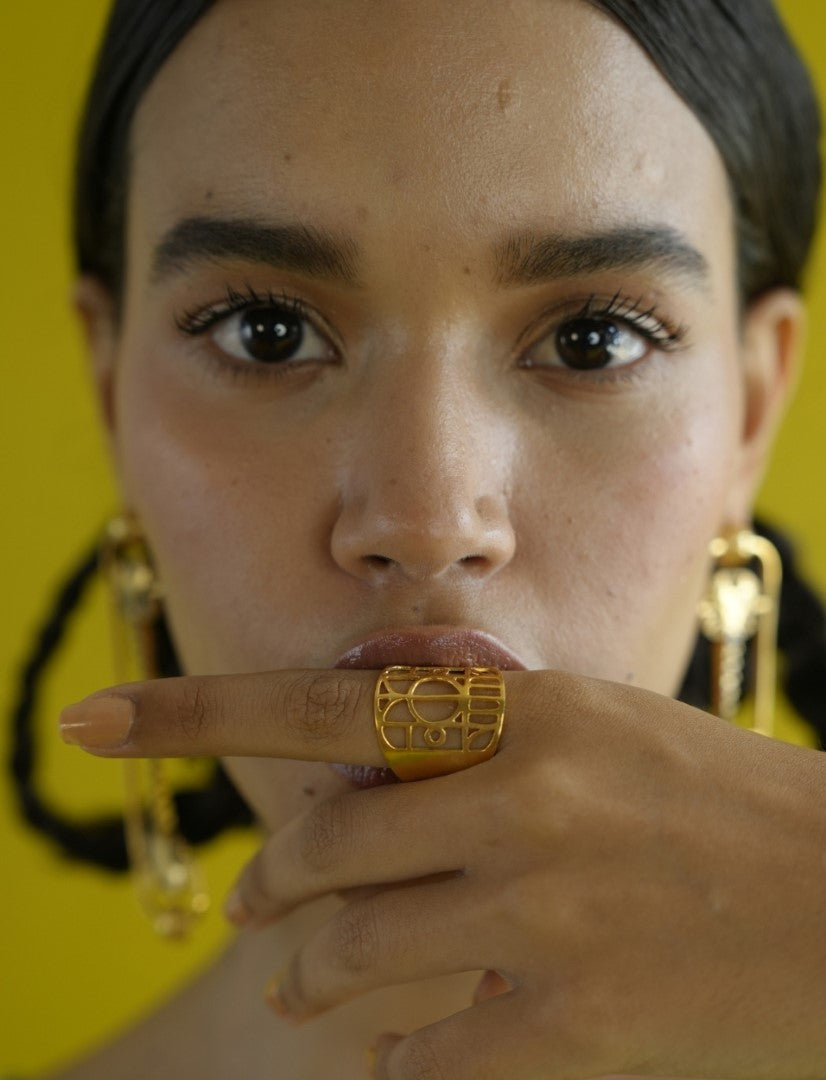 Close-up of woman with dark hair wearing gold geometric statement ring and matching earrings against yellow background