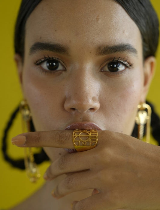Close-up of woman with dark hair wearing gold geometric statement ring and matching earrings against yellow background