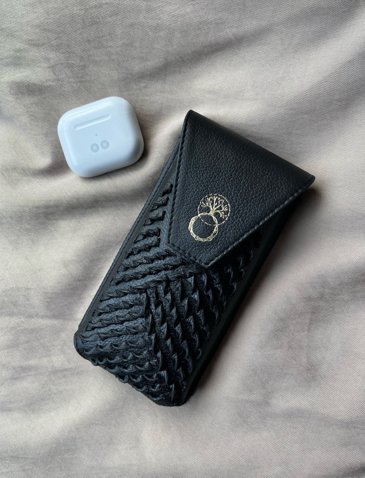 Black upcycled leather iPhone sling with woven design and gold tree logo, next to white wireless earbuds case