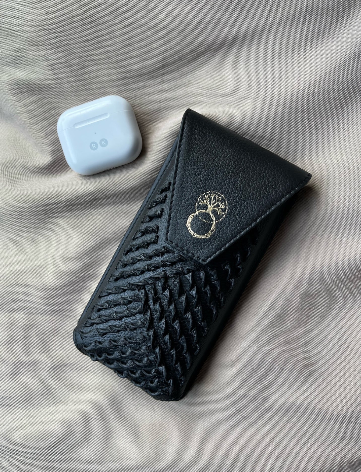 Black upcycled leather iPhone sling with woven design and gold tree logo, next to white wireless earbuds case
