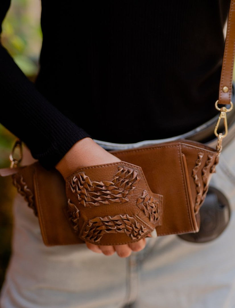 Hand holding brown leather wristlet clutch with woven details, worn crossbody with gold hardware