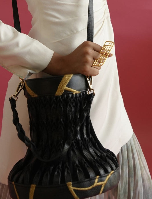 Hand holding gold geometric metal ring with black leather and woven handbag against maroon background