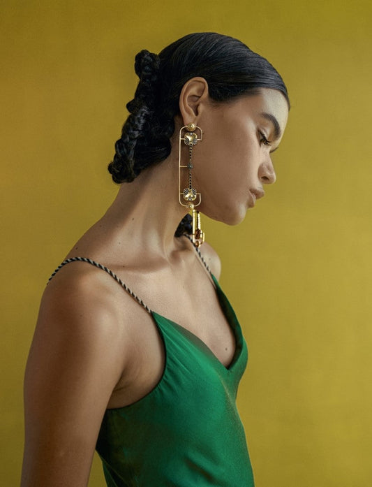 Woman with braided hair wearing a green satin dress and oversized geometric gold earrings against a mustard yellow background