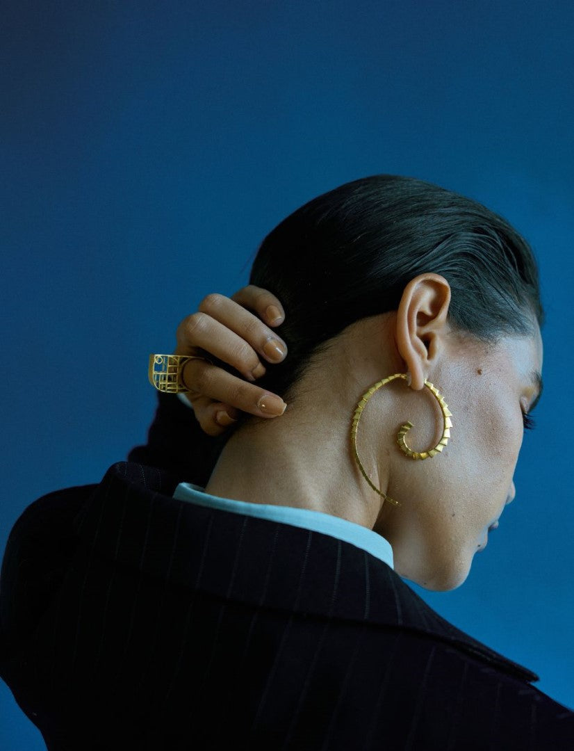 Side profile of woman with slicked black hair wearing large gold spiral earrings and a gold ring against blue background