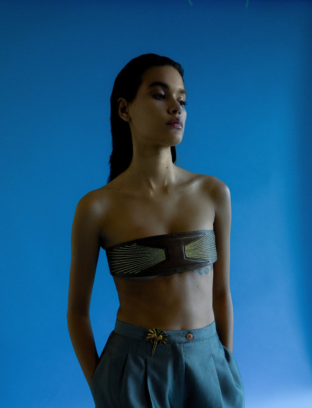Model wearing a timeless tan leather upcycled waistbelt with golden embroidery and high-waisted blue pants against a blue background