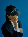 Person wearing black leather visor hat with gold embroidery, dark pinstripe blazer, white shirt, gold earrings, and ring against blue background