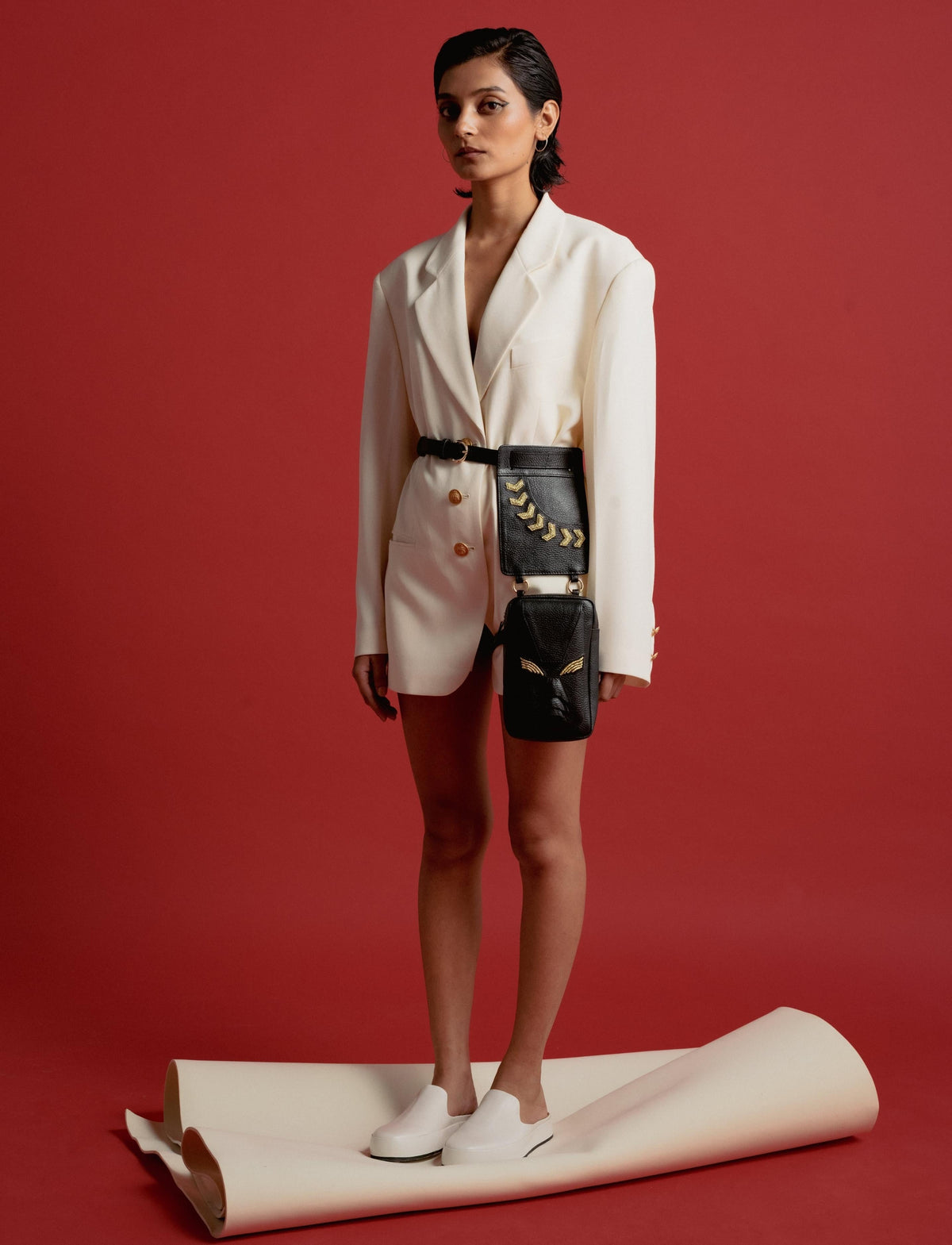 Woman in white oversized blazer dress with black belt bag featuring gold details on red background