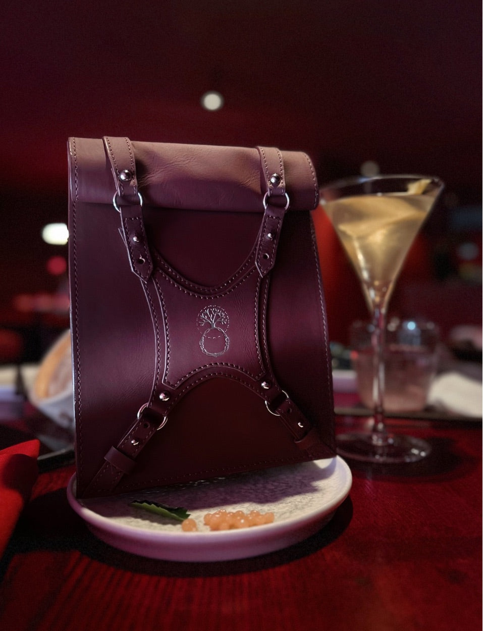 Dark red leather handbag with buckles on a plate beside a martini glass on red table