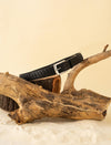 Black woven leather belt with silver buckle displayed on rustic wooden logs and beige sand