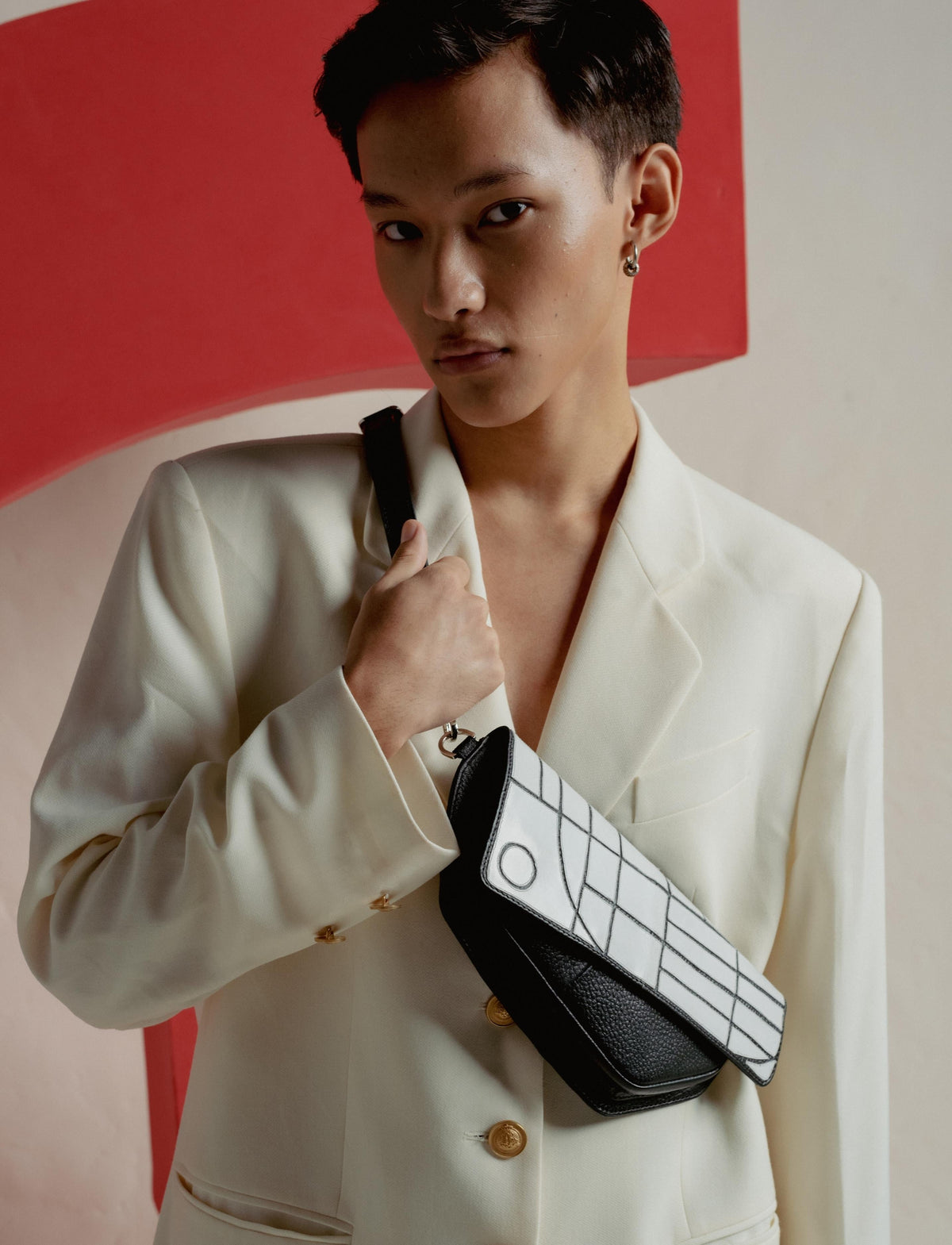 Stylish young man wearing cream blazer and holding black and white patterned crossbody bag against red and beige backdrop