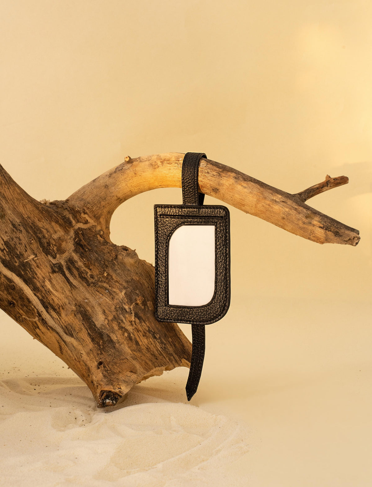 Black leather luggage tag hanging on rustic driftwood with sandy beige background
