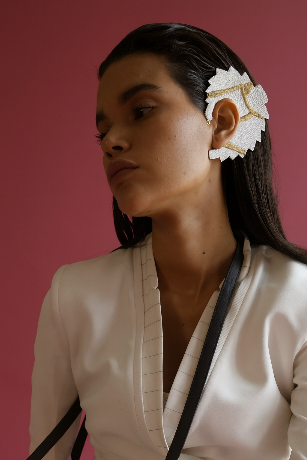 Woman with sleek dark hair wearing white leather and gold embroidered leaf-shaped adjustable ear cuff against pink background