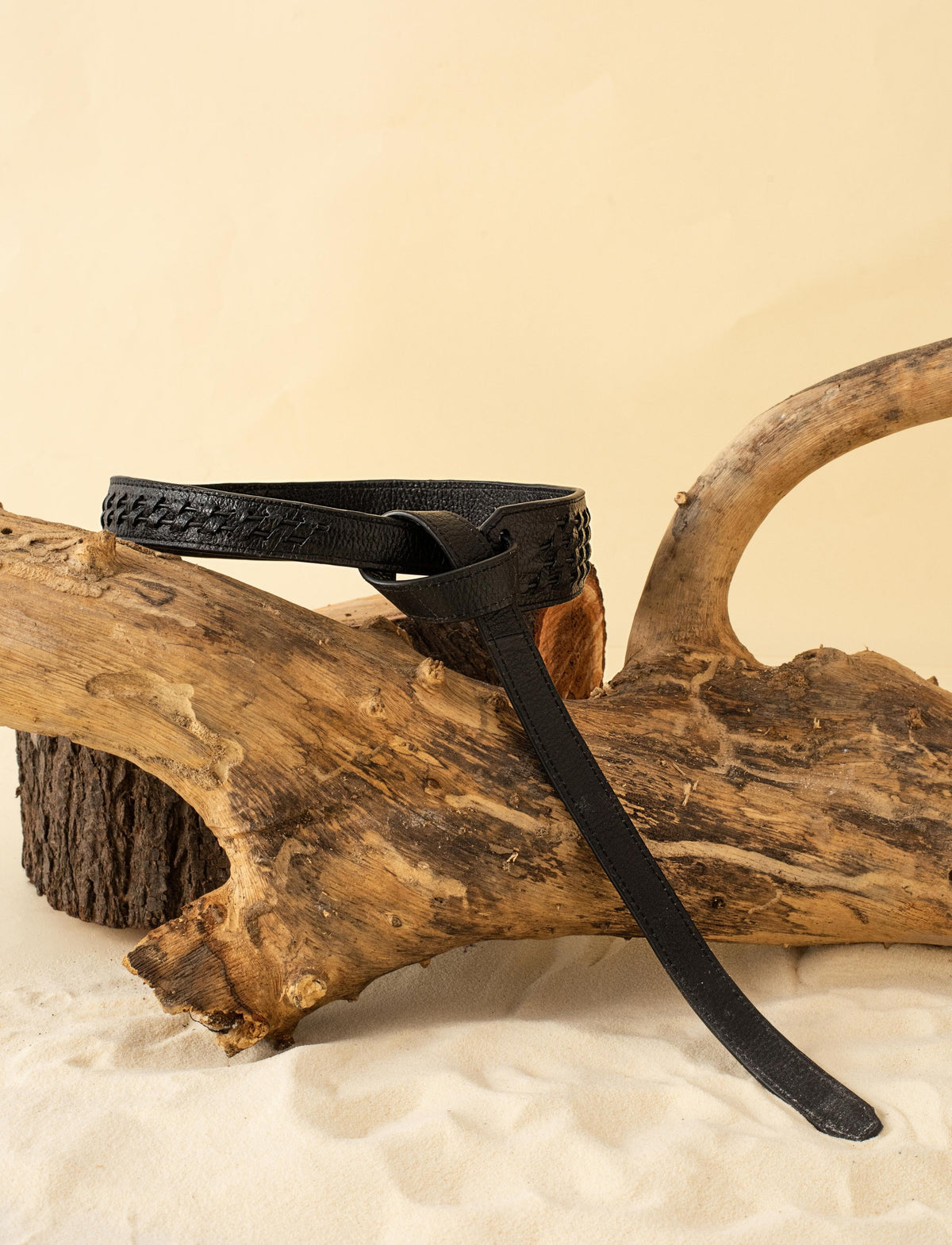 Black leather belt with woven details draped over natural driftwood on sand against beige background