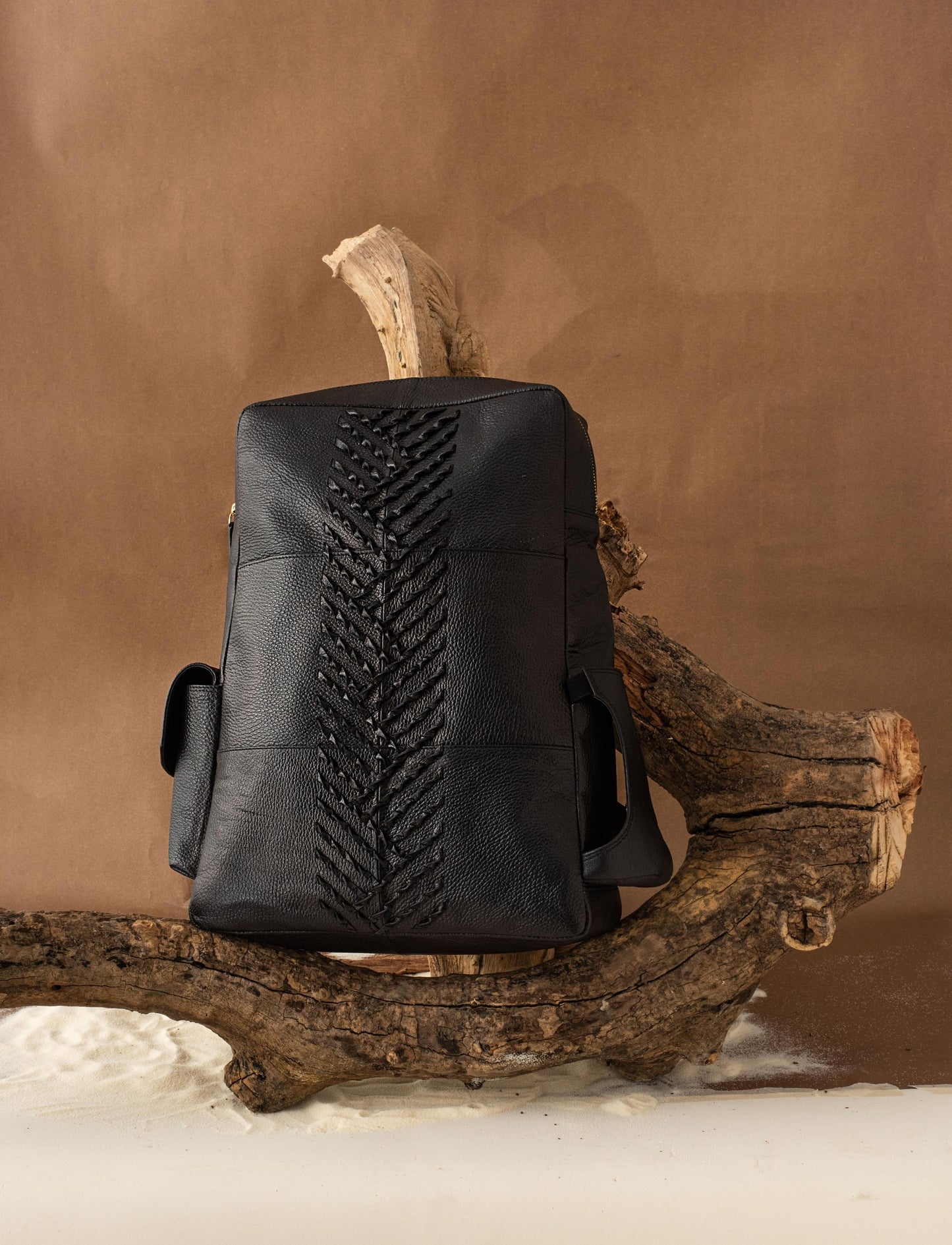 Designer Black leather backpack with braided detailing displayed on a rustic wooden branch against brown background