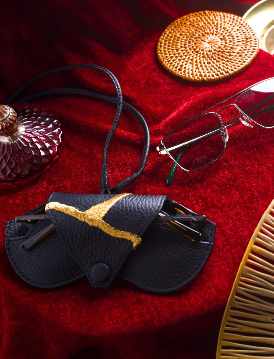 Black leather eyeglass case with gold detail, eyeglasses, rattan coaster, and vintage perfume bottle on red velvet