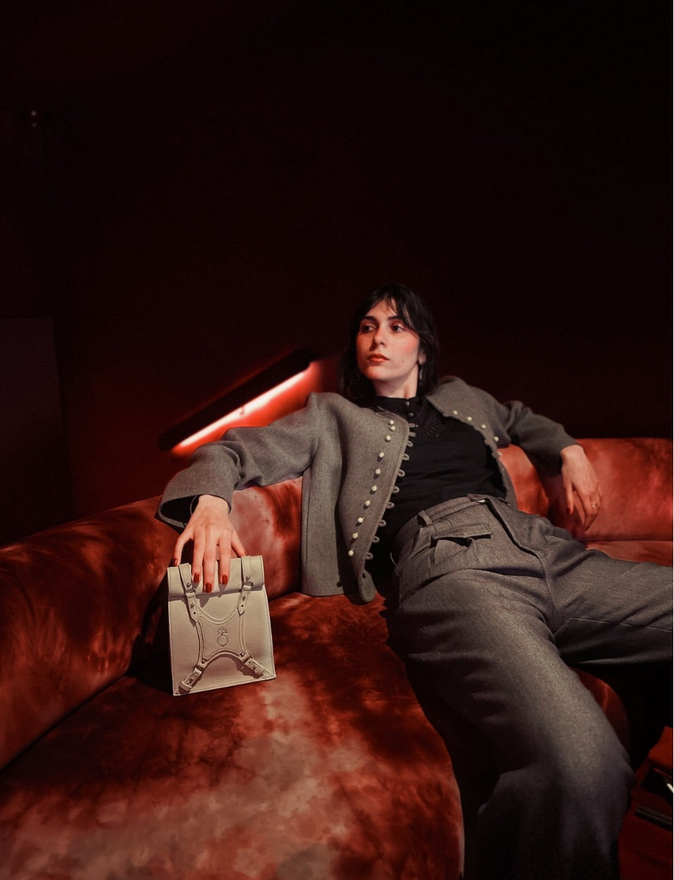Woman in gray jacket and pants lounging on red velvet sofa holding beige handbag in dimly lit room