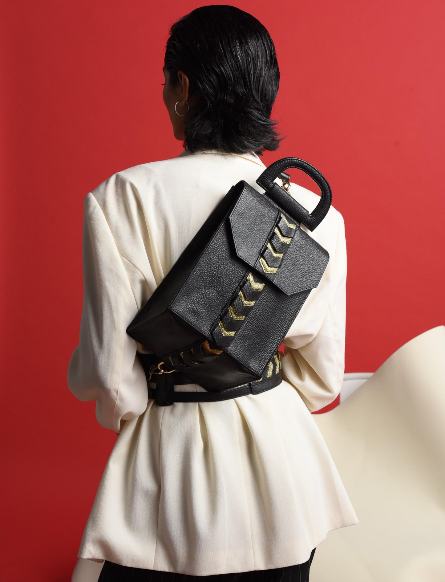 Back view of woman in white blazer wearing black leather backpack with gold arrow detail against red background