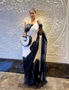 Woman wearing black and white saree with floral embroidery, holding crescent-shaped handbag, standing by a decorative wall