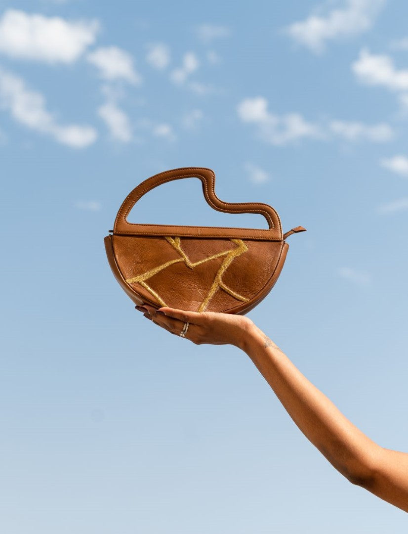 Hand holding brown leather handbag with yellow stitched design against blue sky