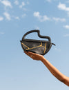 Hand holding black leather semi-circle handbag with gold stitch detail against blue sky