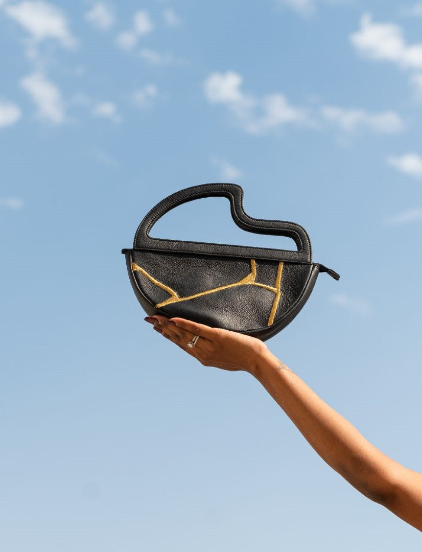 Hand holding black leather semi-circle handbag with gold stitch detail against blue sky