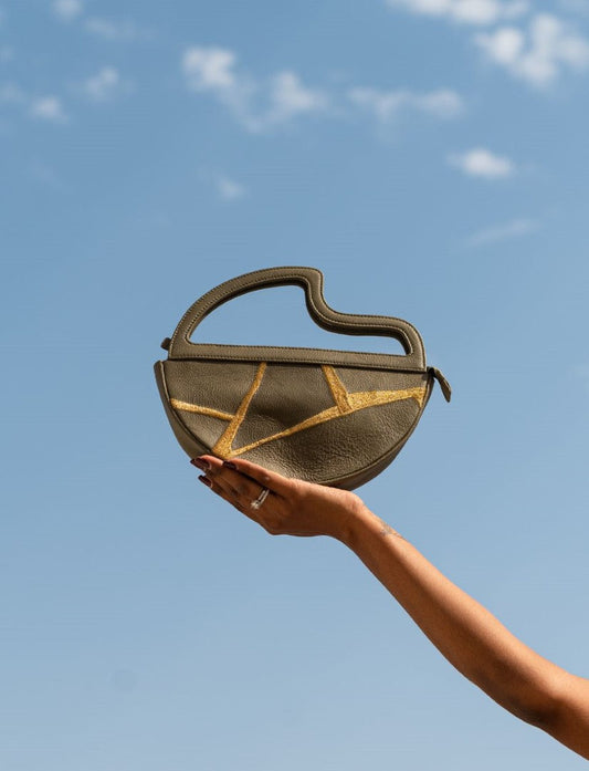 Hand holding olive green leather handbag with gold geometric accents against blue sky