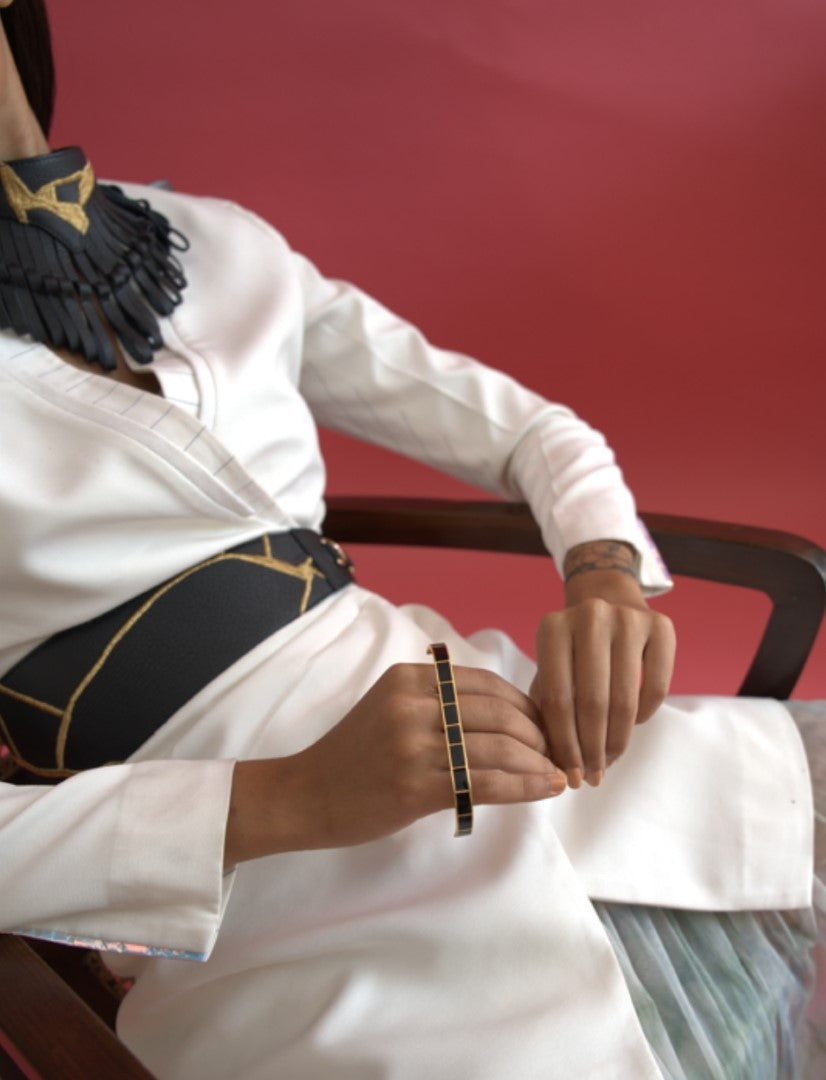 Person wearing white outfit and black wide belt sitting on wooden chair with black and gold metal multifinger ring and black statement necklace