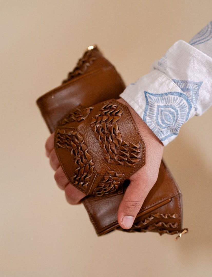 Hand holding a handcrafted tan leather wristlet with braided detailing against beige background